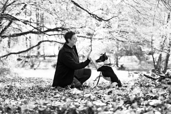 Eine Frau kniet neben einem Hund in einer herbstlichen Landschaft mit Laub. Ein Mann kniet neben einem Hund in einer herbstlichen Landschaft mit Laub.