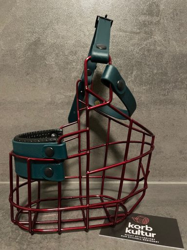 Rotem Drahtkorb mit blauer Lederapplikation für Hunde, auf grauem Hintergrund.